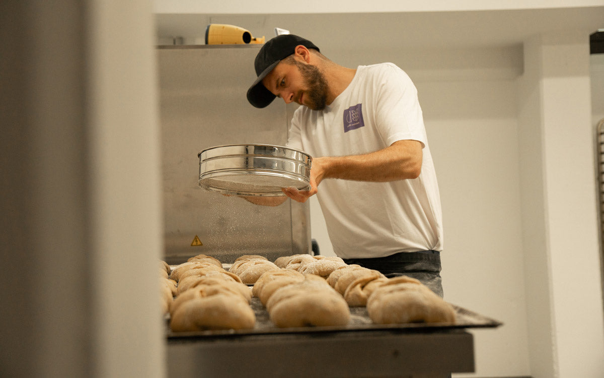 Bäcker bestäubt vor dem Backen Brötchen mit Mehl. Handgemachte Backwaren online kaufen bei Backverliebt.com